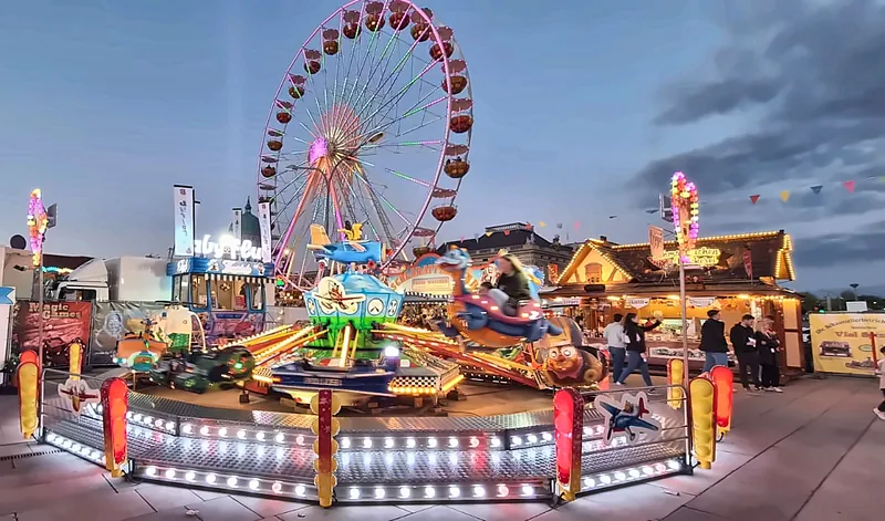 Der Babyflug in der Abenddämmerung: Kinderkarussell mit Flugzeugen und Lichterkranz, dahinter ein Riesenrad.