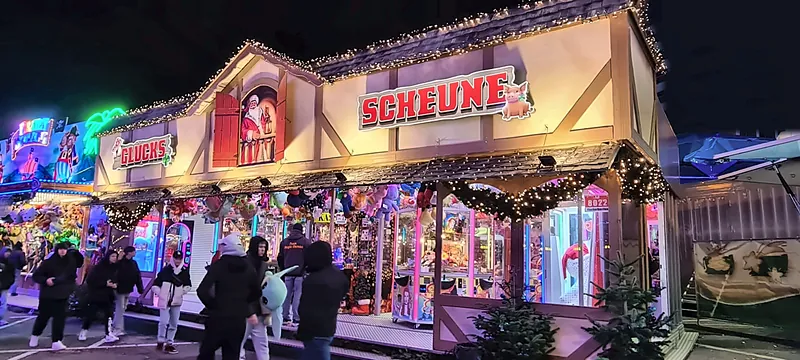 Die Glücks Scheune abends auf dem Weihnachtsmarkt, Lichterketten am Dach, viele Besucher davor.